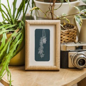 Hand carved lino block print of a hanging plant in a macrame holder on handmade paper in wooden frame surrounded by house plants and camera